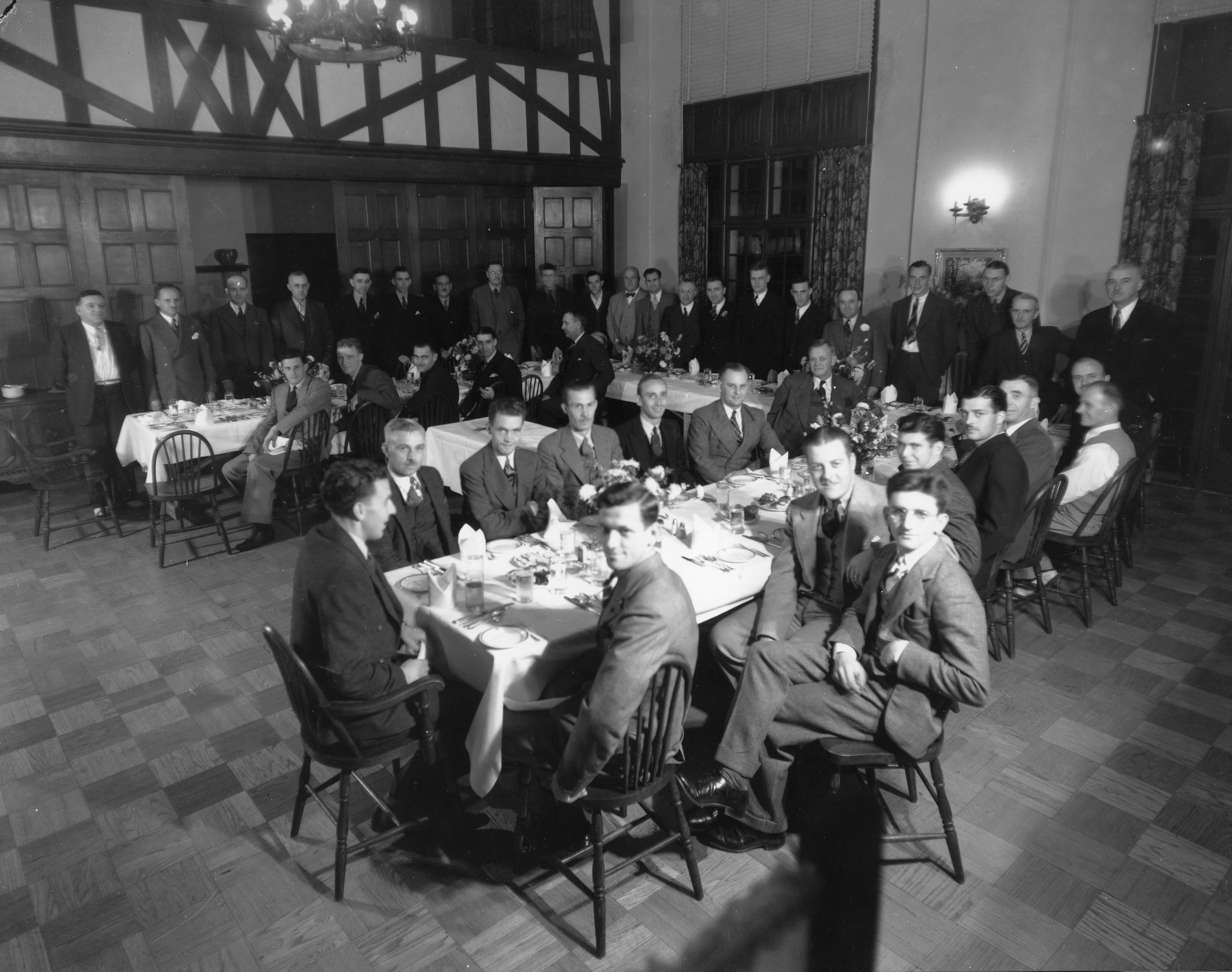 Historical black and white photograph depicting a group of distinguished men in formal attire seated around tables at a large banquet hall, seated and standing, staring directly into the camera, suggesting a formal event or gathering from an earlier era. It is almost as if we are interrupting an important meeting.