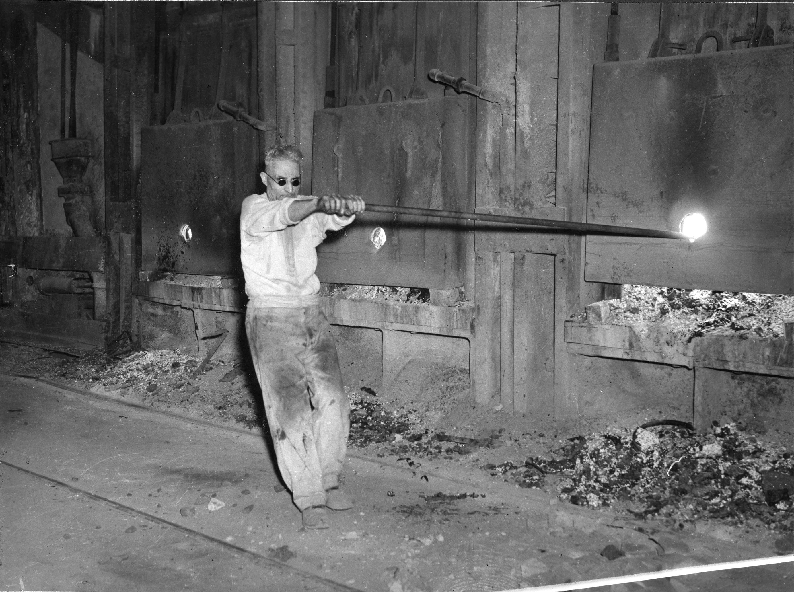 Vintage black and white photo of a Hamilton steelworker in circular fashion forward protective glasses, with rolled up shirt sleeves and very minimal protective gear, holding a long rod in an industrial furnace setting, with molten material and machinery in the background.