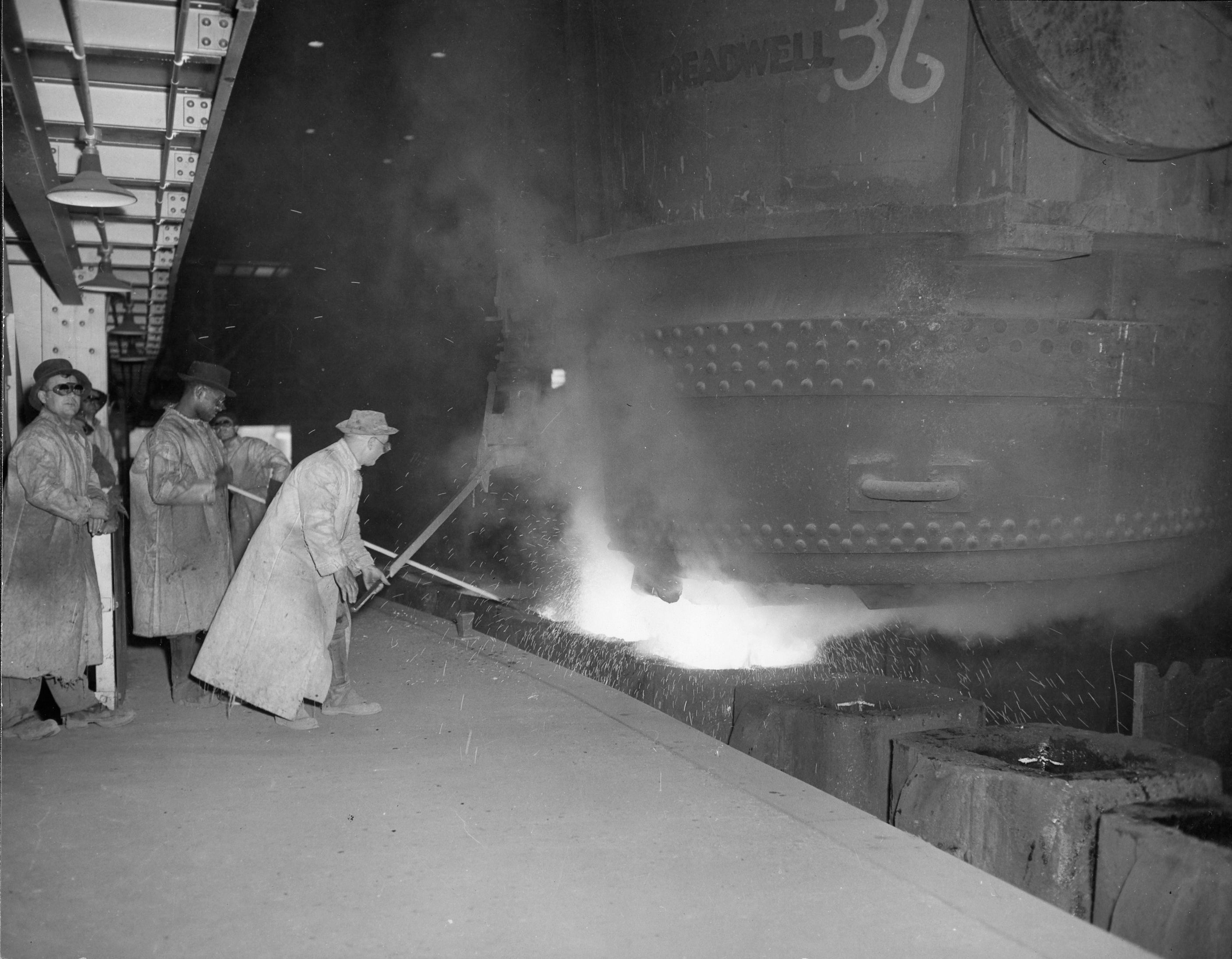 Historical black and white photo of 5 Hamilton steelworkers is long trench coats, fedora hats and protective round "sunglasses" pouring molten metal in an industrial setting, with the number 36 visible on equipment, highlighting the steel-making process.