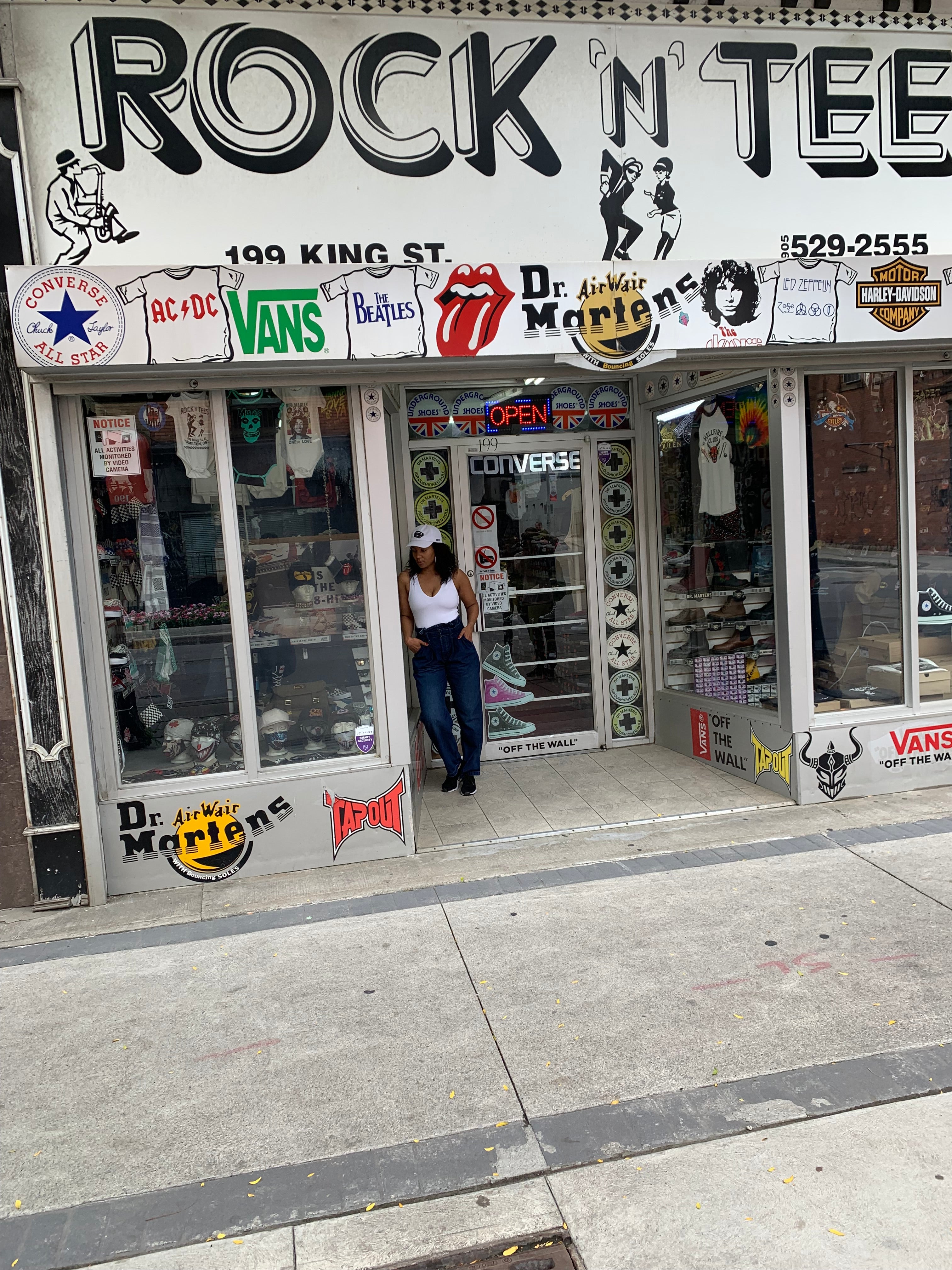 A young black female in a white RUBBERBAND SOVEREIGN GOLF CAP, leans against a store front of the iconic 'ROCK N' TEE' store, in downtown Hamilton on 199 King St., featuring a display of iconic brand logos such as Vans, The Beatles, AC/DC, and Dr. Martens. The storefront showcases a variety of merchandise visible through the glass windows