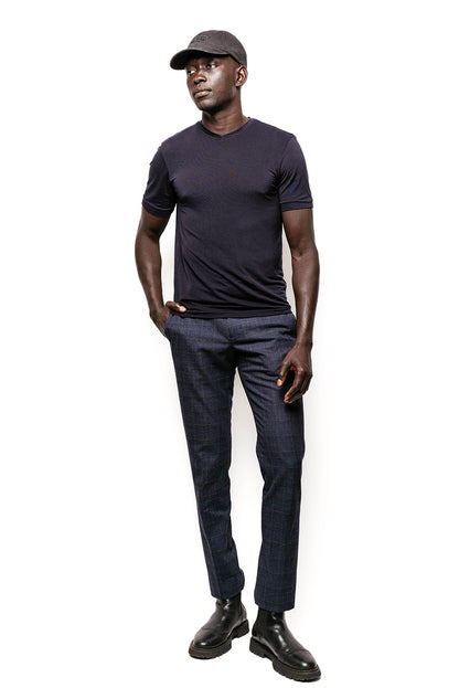 A male model standing against a white background. Wearing a dark, textured form fitting T-shirt and a pair of dark denim tailored trousers that feature a subtle checked pattern. Contributing to a sleek and polished look, he compliments the outfit with a black RSG cap, which adds a refined touch to the overall ensemble, paired with black leather boots that have a thick sole, giving a sturdy and robust appearance. His pose and the attire suggest a blend of casual sophistication with an urban edge.