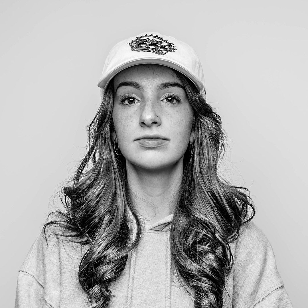 A young woman in a monochrome setting, with long curly red hair is shown in profile, in a grey hoodie sweater wearing a white, six-panel cap, crafted from 100% cotton. The front panel features the Rubberband Sovereign detailed "crowned" logo embroidered in black. On the left side of the cap, "MMXXIII" is neatly embroidered in black, signifying the year 2023 in Roman numerals. The unstructured, low-profile design gives the cap a relaxed and comfortable fit.