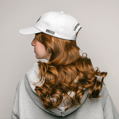 The side profile of a young female model with long curly red hair, hooped earrings and pierced diamond nose ring, wearing a white, one-size-fits-all 6-panel cap, made of 100% cotton. On the left side of the cap, "MMXXIII" is neatly embroidered in black, signifying the year 2023 in Roman numerals. The back of the cap features the brand name "RUBBERBAND SOVEREIGN" also embroidered in black, situated above the cap's adjustable opening and RSG woven white and black loop label.