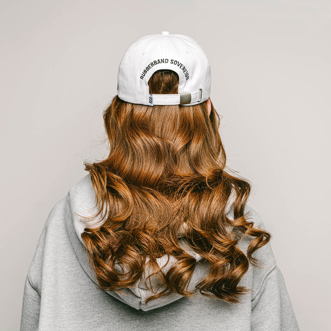 The back profile of a young female model with long curly red hair, wearing a white , one-size-fits-all 6-panel cap, made of 100% cotton. The back of the cap features the brand name "RUBBERBAND SOVEREIGN" embroidered in black, situated above the cap's adjustable opening with a RSG woven white and black loop label. The cap is secured with a metal clasp closure, allowing for a customizable fit and the unstructured, low-profile design gives the cap a relaxed and comfortable fit.