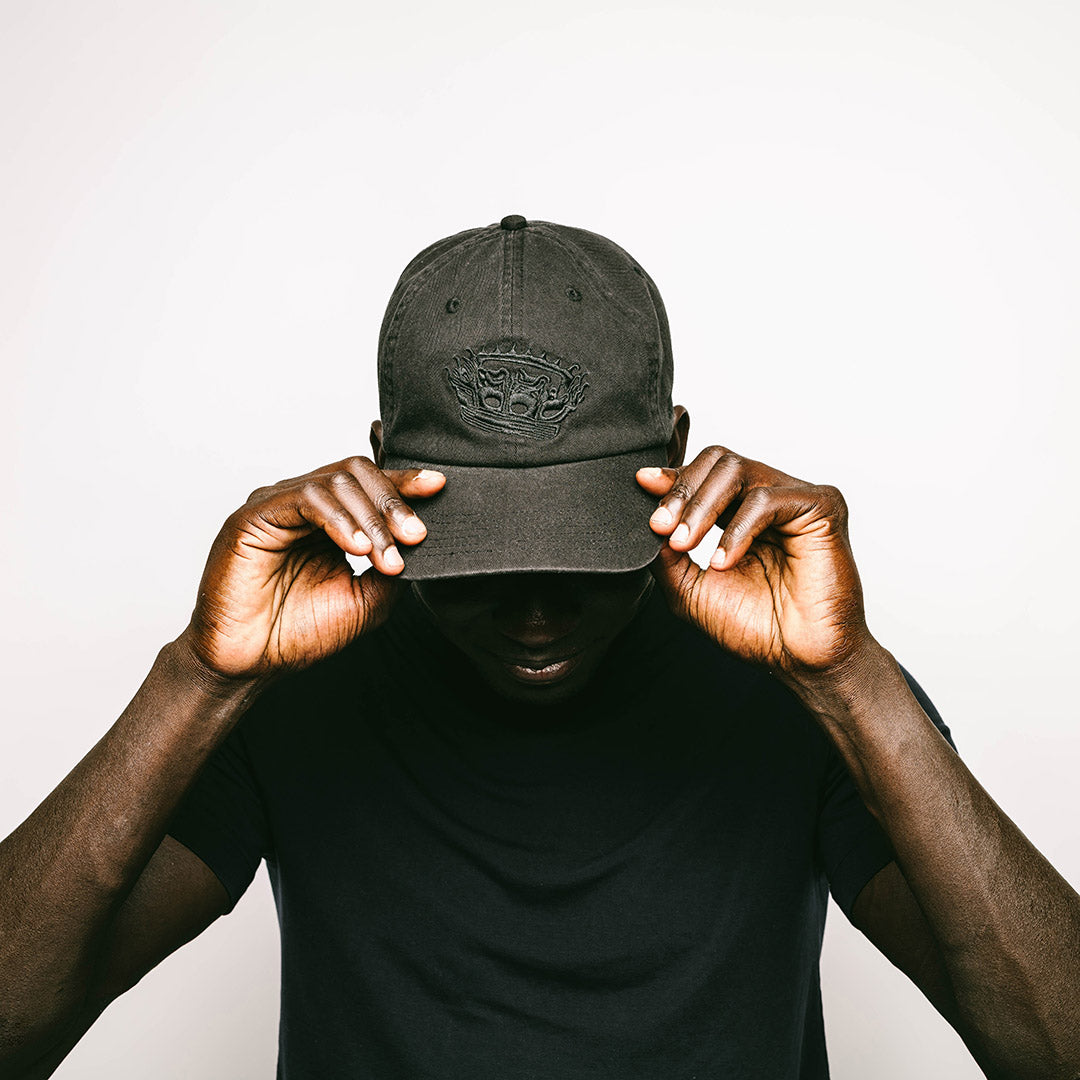A confident male model adjusts a black, cotton 6-panel cap with subtle, tonal black embroidery of the RSG detailed crown, featuring an adjustable metal clasp for a custom fit and a relaxed, unstructured silhouette.