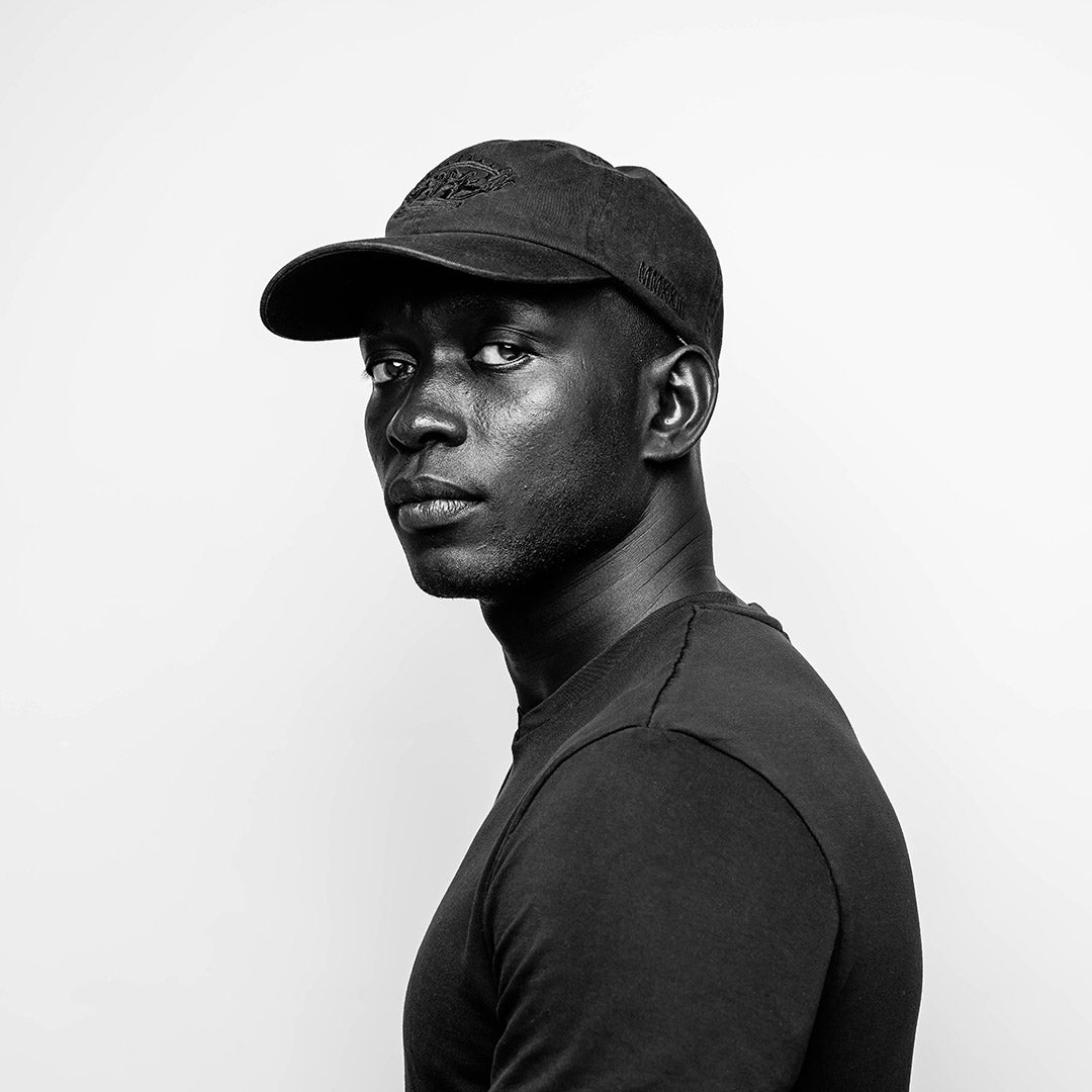 A monochromatic portrait of a young black man wearing a black on black embroidered RUBBERBAND SOVEREIGN ball cap and t-shirt, gazing to the side with a thoughtful expression, highlighting subtle textures and contrasts