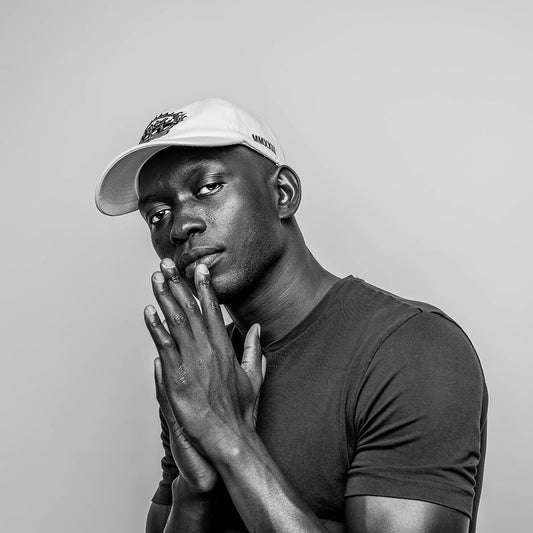 A male model in a monochrome setting, wearing a white, RSG 6-panel cotton cap. On the left side of the cap, "MMXXIII" is embroidered in black. Dressed in a close-fitting, dark crew neck T-shirt. The model's pose is striking, with his hands clasped together in front of him in a thoughtful and intentional gesture, drawing attention to the elegance of his posture and the thoughtful expression on his face. His gaze is direct and contemplative, inviting a connection with the viewer. 