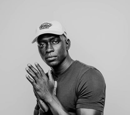 A male model in a monochrome setting, wearing a white, 6-panel cotton cap. On the front panel  of the cap, the RSG "Crown" is embroidered in black. Dressed in a close-fitting, dark crew neck T-shirt. The model's pose is striking, with his hands clasped together in front of him in a thoughtful and intentional gesture, drawing attention to the elegance of his posture and the thoughtful expression on his face. His gaze is direct and contemplative, inviting a connection with the viewer.