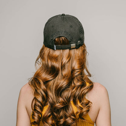 The back profile of a young female model with long curly red hair, wearing a black , one-size-fits-all 6-panel cap, made of 100% cotton. The back of the cap features the brand name "RUBBERBAND SOVEREIGN" embroidered in black, situated above the cap's adjustable opening with a RSG woven white and black loop label. The cap is secured with a metal clasp closure, allowing for a customizable fit and the unstructured, low-profile design gives the cap a relaxed and comfortable fit.