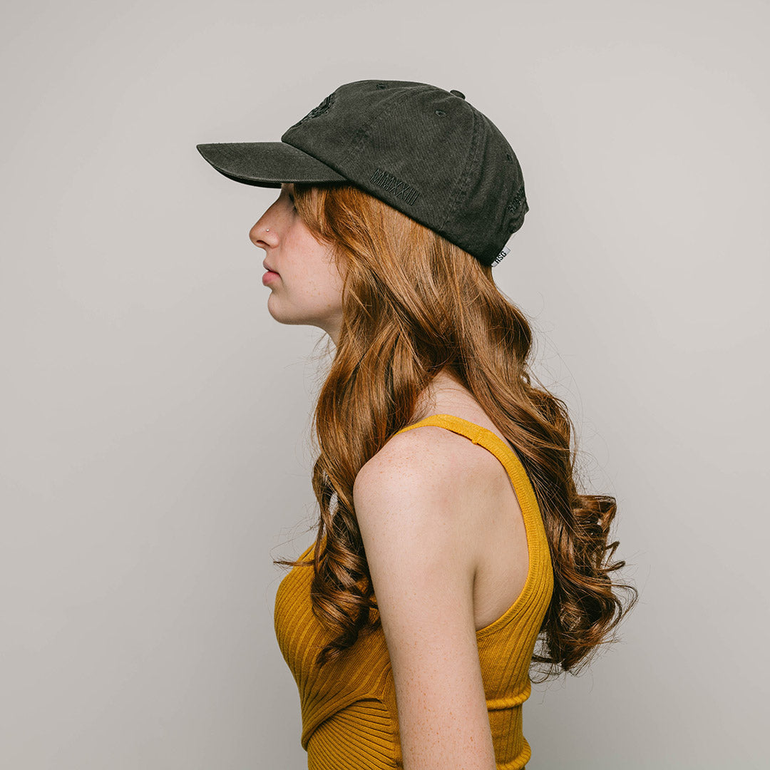  The side profile of a young female model with long curly red hair, wearing a black, one-size-fits-all 6-panel cap, made of 100% cotton. On the left side of the cap, "MMXXIII" is neatly embroidered in black, signifying the year 2023 in Roman numerals. The back of the cap features the brand name "RUBBERBAND SOVEREIGN" also embroidered in black, situated above the cap's adjustable opening. Just below this text is the RSG woven loop label.