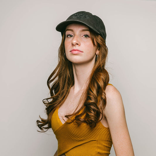 A young female with long, wavy auburn hair, wearing a black, 6-panel cap adorned with the detailed tonal embroidery of the RSG "crowned" logo . The cap’s design is understated yet elegant, complementing her mustard yellow top and confident, poised demeanor.