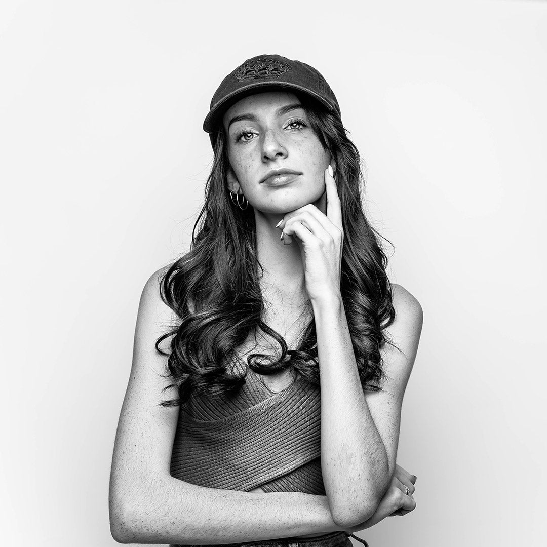 A monochrome portrait of a teenage girl with long curly hair, wearing a gold sleeveless top and a black RUBBERBAND SOVEREIGN ball cap with intricate black embroidery of the RSG crown logo, thoughtfully touching her cheek with a finger.